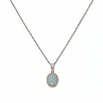 An Ethereal Opal with White Cubic Zirconia Halo Pendant Necklace by Skōsh Jewelry, featuring an oval iridescent opal framed by a halo of small clear stones on a fine sterling silver chain, displayed against a white background.