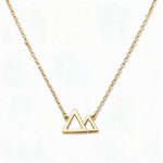 A Mountains and Valleys Necklace by Skōsh Jewelry, featuring an open outline of two mountain peaks set on a fine gold chain, displayed against a white background.