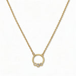 A Trio White Cubic Zirconia Accent Circle Necklace by Skōsh Jewelry, featuring a small open circle accented with three clear stones on a fine gold chain, displayed against a white background.