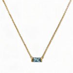 A March Birthstone Light Sapphire Cubic Zirconia Baguette Necklace by Skōsh Jewelry, featuring a slim rectangular light blue baguette stone set horizontally on a fine gold chain, displayed against a white background.