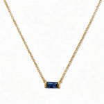 A September Birthstone Sapphire Cubic Zirconia Baguette Necklace by Skōsh Jewelry, featuring a slim rectangular deep blue baguette stone set horizontally on a fine gold chain, displayed against a white background.