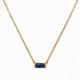 A September Birthstone Sapphire Cubic Zirconia Baguette Necklace by Skōsh Jewelry, featuring a slim rectangular deep blue baguette stone set horizontally on a fine gold chain, displayed against a white background.