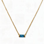 A December Birthstone Aqua Cubic Zirconia Baguette Necklace by Skōsh Jewelry, featuring a slim rectangular blue baguette stone set horizontally on a fine gold chain, displayed against a white background.