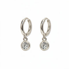 A pair of Bezel Set White Cubic Zirconia Drop Huggie Hoop Earrings by Skōsh Jewelry, featuring small sterling silver huggie hoops, each with a round bezel-set clear stone dangling below, displayed against a white background.