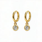 A pair of Bezel Set White Cubic Zirconia Drop Huggie Hoop Earrings by Skōsh Jewelry, featuring small gold huggie hoops, each with a round bezel-set clear stone dangling below, displayed against a white background.