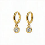 A pair of Bezel Set White Cubic Zirconia Drop Huggie Hoop Earrings by Skōsh Jewelry, featuring small gold huggie hoops, each with a round bezel-set clear stone dangling below, displayed against a white background.