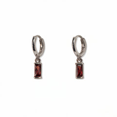 A pair of January Birthstone Garnet Cubic Zirconia Baguette Earrings by Skōsh Jewelry, featuring small sterling silver huggie hoops, each with a rectangular deep red baguette stone dangling below, displayed against a white background.