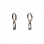 A pair of April Birthstone White Cubic Zirconia Baguette Earrings by Skōsh Jewelry, featuring small sterling silver huggie hoops, each with a rectangular clear baguette stone dangling below, displayed against a white background.