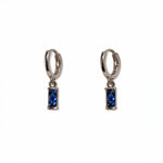 A pair of September Birthstone Sapphire Cubic Zirconia Baguette Earrings by Skōsh Jewelry, featuring small sterling silver huggie hoops, each with a rectangular deep blue baguette stone dangling below, displayed against a white background.
