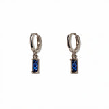 A pair of September Birthstone Sapphire Cubic Zirconia Baguette Earrings by Skōsh Jewelry, featuring small sterling silver huggie hoops, each with a rectangular deep blue baguette stone dangling below, displayed against a white background.
