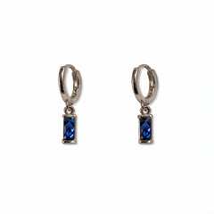 A pair of September Birthstone Sapphire Cubic Zirconia Baguette Earrings by Skōsh Jewelry, featuring small sterling silver huggie hoops, each with a rectangular deep blue baguette stone dangling below, displayed against a white background.