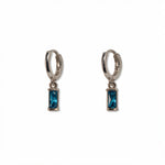 A pair of December Birthstone Aqua Cubic Zirconia Baguette Earrings by Skōsh Jewelry, featuring small sterling silver huggie hoops, each with a rectangular blue baguette stone dangling below, displayed against a white background.
