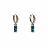 A pair of December Birthstone Aqua Cubic Zirconia Baguette Earrings by Skōsh Jewelry, featuring small sterling silver huggie hoops, each with a rectangular blue baguette stone dangling below, displayed against a white background.