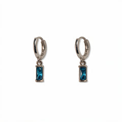 A pair of December Birthstone Aqua Cubic Zirconia Baguette Earrings by Skōsh Jewelry, featuring small sterling silver huggie hoops, each with a rectangular blue baguette stone dangling below, displayed against a white background.