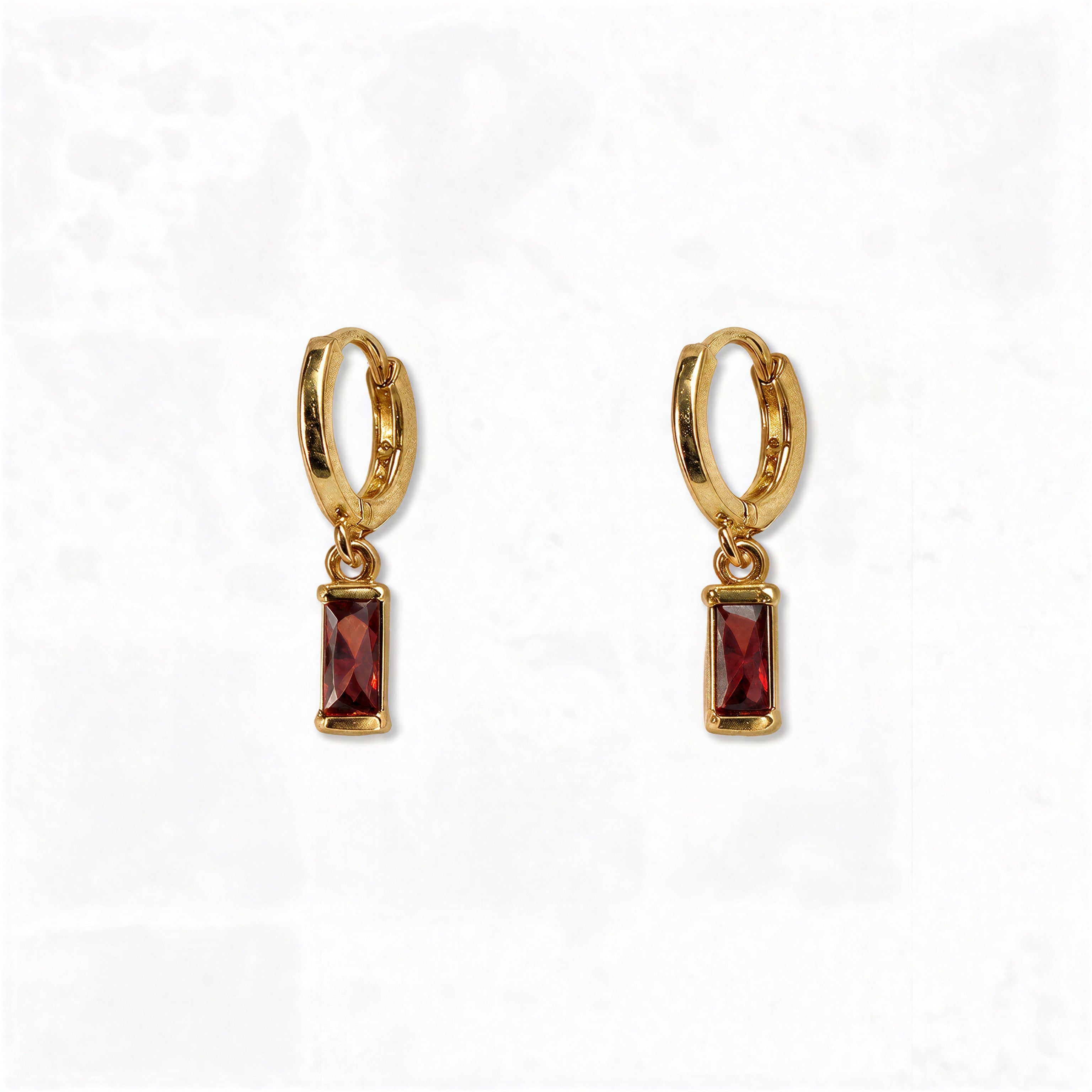 A pair of January Birthstone Garnet Cubic Zirconia Baguette Earrings by Skōsh Jewelry, featuring small gold huggie hoops, each with a rectangular deep red baguette stone dangling below, displayed against a white background.