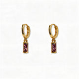 A pair of February Birthstone Amethyst Cubic Zirconia Baguette Earrings by Skōsh Jewelry, featuring small gold huggie hoops, each with a rectangular purple baguette stone dangling below, displayed against a white background.