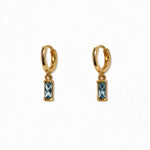 A pair of March Birthstone Light Sapphire Cubic Zirconia Baguette Earrings by Skōsh Jewelry, featuring small gold huggie hoops, each with a rectangular light blue baguette stone dangling below, displayed against a white background.