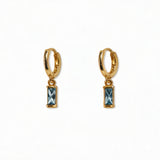 A pair of March Birthstone Light Sapphire Cubic Zirconia Baguette Earrings by Skōsh Jewelry, featuring small gold huggie hoops, each with a rectangular light blue baguette stone dangling below, displayed against a white background.