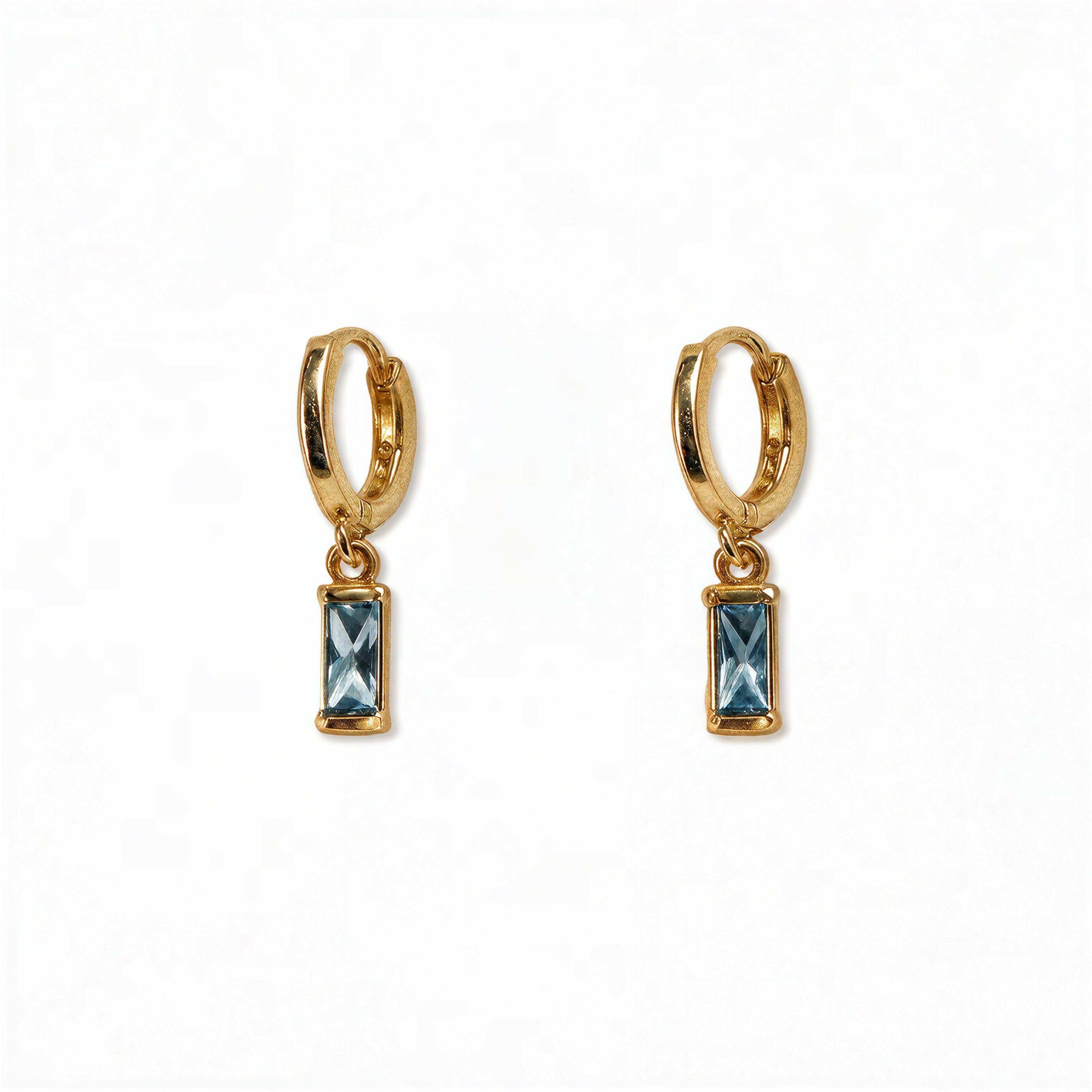 A pair of March Birthstone Light Sapphire Cubic Zirconia Baguette Earrings by Skōsh Jewelry, featuring small gold huggie hoops, each with a rectangular light blue baguette stone dangling below, displayed against a white background.