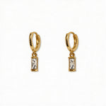 A pair of April Birthstone White Cubic Zirconia Baguette Earrings by Skōsh Jewelry, featuring small gold huggie hoops, each with a rectangular clear baguette stone dangling below, displayed against a white background.