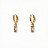 A pair of April Birthstone White Cubic Zirconia Baguette Earrings by Skōsh Jewelry, featuring small gold huggie hoops, each with a rectangular clear baguette stone dangling below, displayed against a white background.