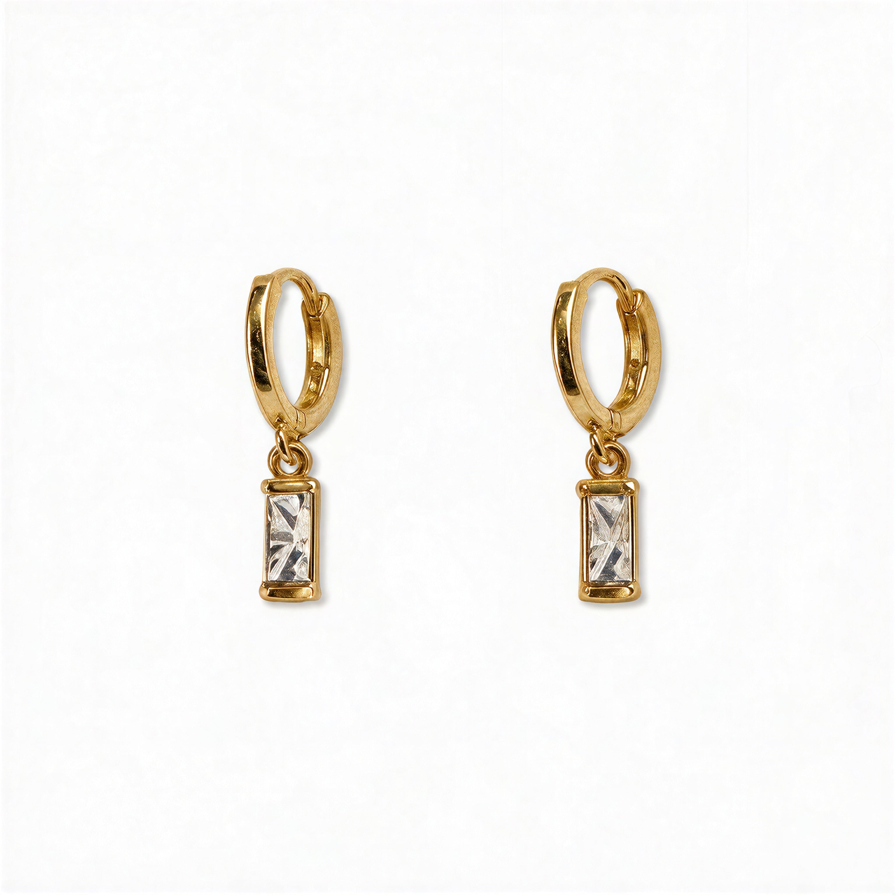 A pair of April Birthstone White Cubic Zirconia Baguette Earrings by Skōsh Jewelry, featuring small gold huggie hoops, each with a rectangular clear baguette stone dangling below, displayed against a white background.