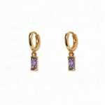 A pair of June Birthstone Lavender Cubic Zirconia Baguette Earrings by Skōsh Jewelry, featuring small gold huggie hoops, each with a rectangular light purple baguette stone dangling below, displayed against a white background.
