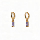 A pair of June Birthstone Lavender Cubic Zirconia Baguette Earrings by Skōsh Jewelry, featuring small gold huggie hoops, each with a rectangular light purple baguette stone dangling below, displayed against a white background.