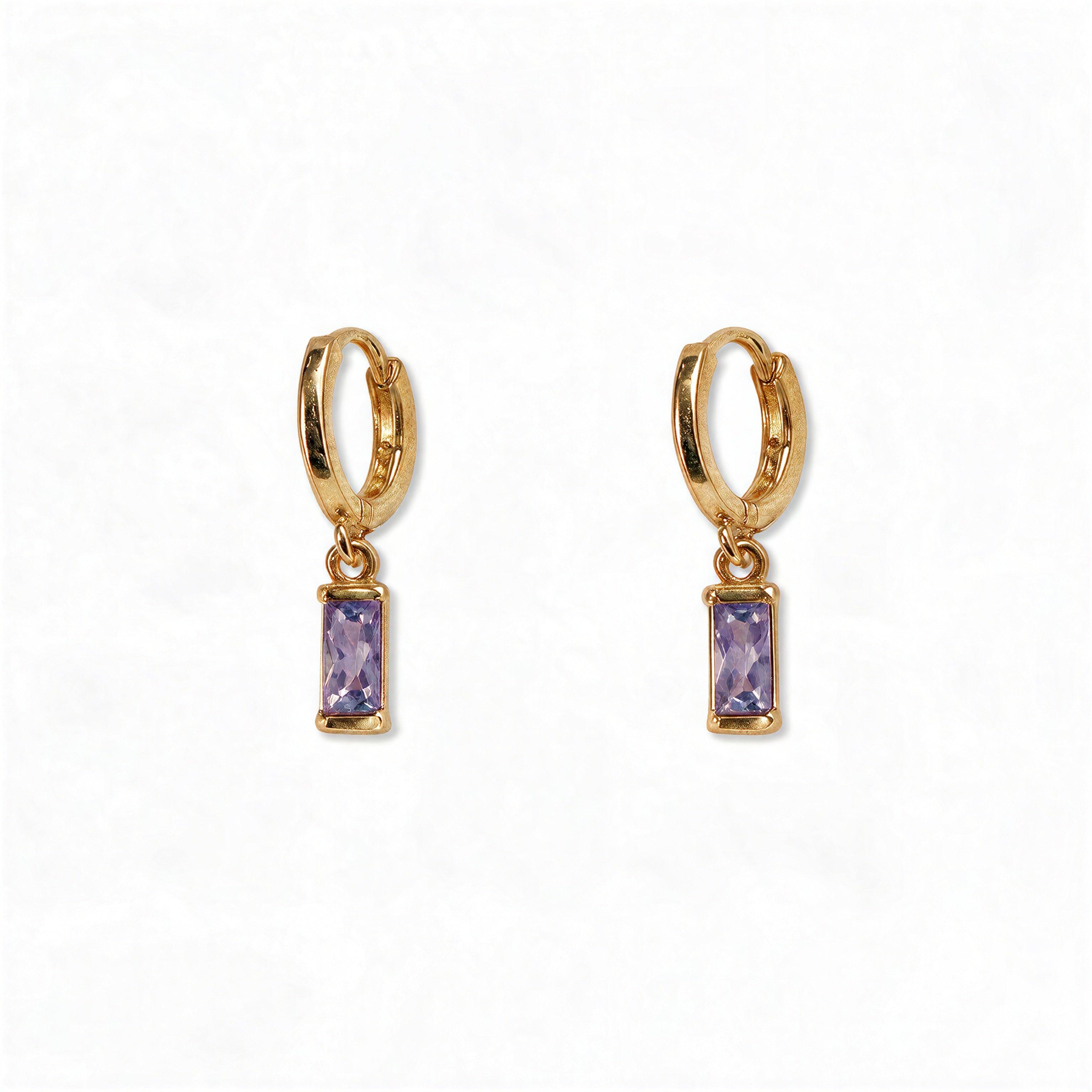 A pair of June Birthstone Lavender Cubic Zirconia Baguette Earrings by Skōsh Jewelry, featuring small gold huggie hoops, each with a rectangular light purple baguette stone dangling below, displayed against a white background.