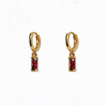 A pair of July Birthstone Ruby Cubic Zirconia Baguette Earrings by Skōsh Jewelry, featuring small gold huggie hoops, each with a rectangular red baguette stone dangling below, displayed against a white background.