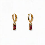 A pair of July Birthstone Ruby Cubic Zirconia Baguette Earrings by Skōsh Jewelry, featuring small gold huggie hoops, each with a rectangular red baguette stone dangling below, displayed against a white background.