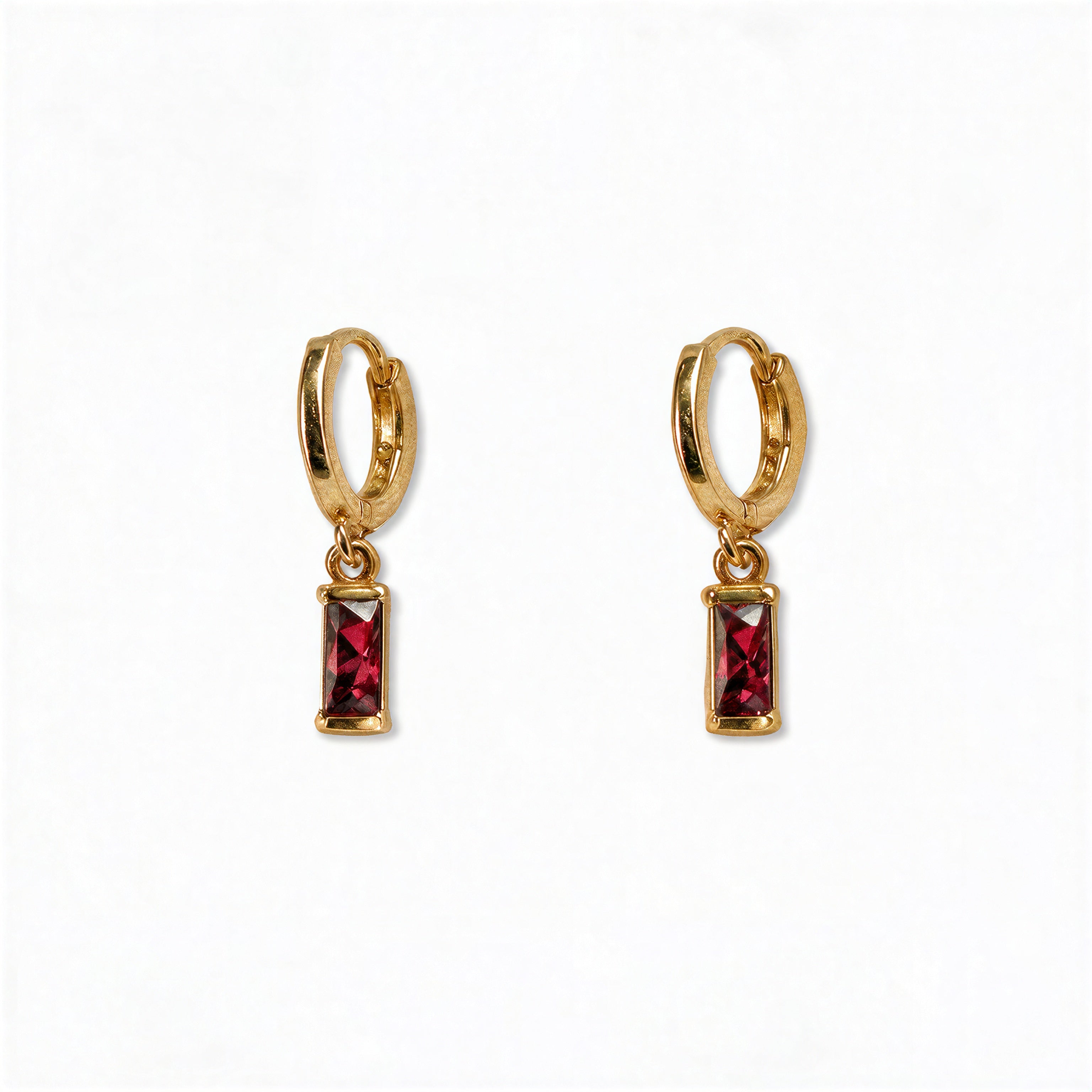 A pair of July Birthstone Ruby Cubic Zirconia Baguette Earrings by Skōsh Jewelry, featuring small gold huggie hoops, each with a rectangular red baguette stone dangling below, displayed against a white background.