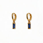 A pair of September Birthstone Sapphire Cubic Zirconia Baguette Earrings by Skōsh Jewelry, featuring small gold huggie hoops, each with a rectangular deep blue baguette stone dangling below, displayed against a white background.