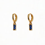 A pair of September Birthstone Sapphire Cubic Zirconia Baguette Earrings by Skōsh Jewelry, featuring small gold huggie hoops, each with a rectangular deep blue baguette stone dangling below, displayed against a white background.