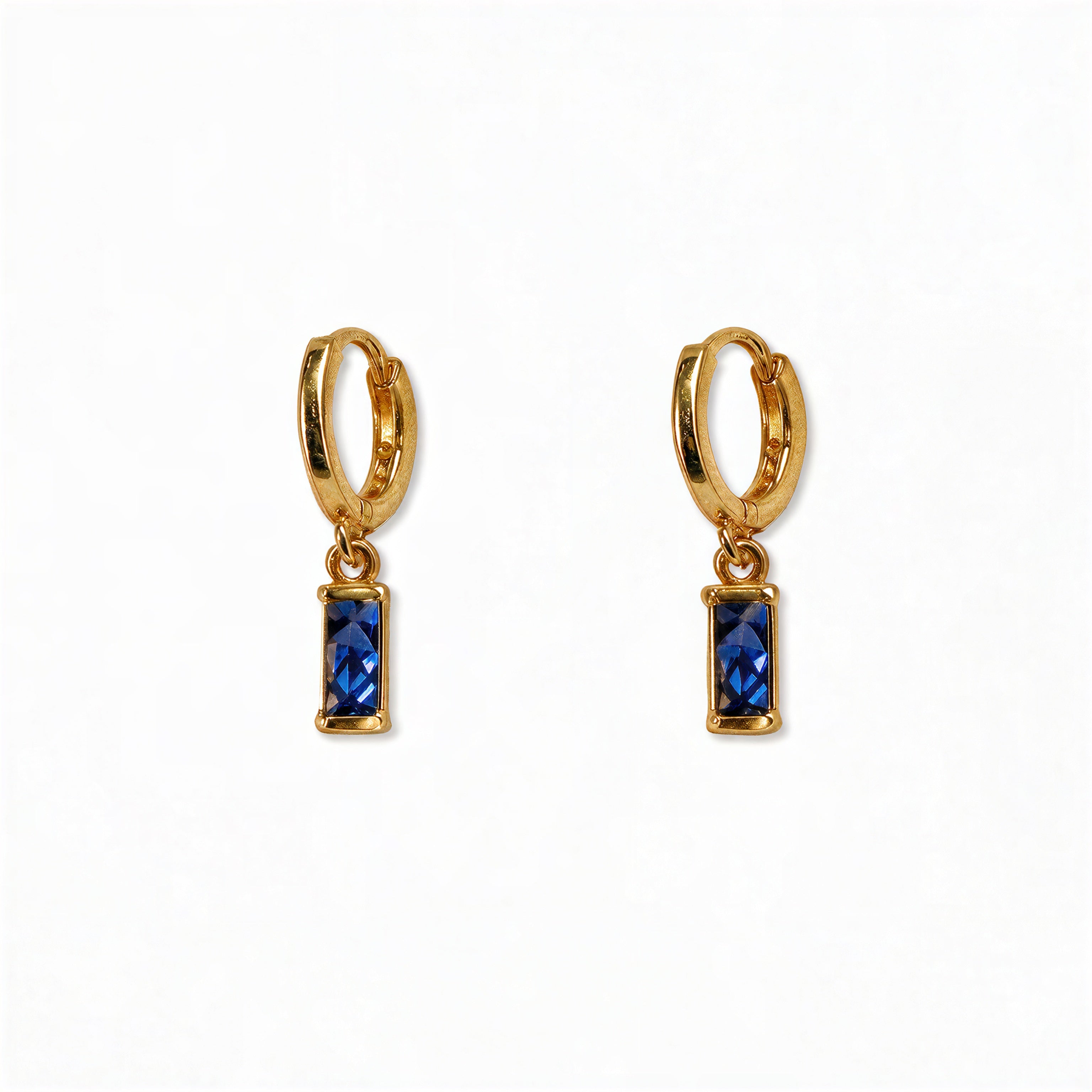 A pair of September Birthstone Sapphire Cubic Zirconia Baguette Earrings by Skōsh Jewelry, featuring small gold huggie hoops, each with a rectangular deep blue baguette stone dangling below, displayed against a white background.