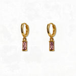 A pair of October Birthstone Pink Cubic Zirconia Baguette Earrings by Skōsh Jewelry, featuring small gold huggie hoops, each with a rectangular pink baguette stone dangling below, displayed against a white background.
