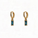 A pair of December Birthstone Aqua Cubic Zirconia Baguette Earrings by Skōsh Jewelry, featuring small gold huggie hoops, each with a rectangular blue baguette stone dangling below, displayed against a white background.