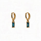 A pair of December Birthstone Aqua Cubic Zirconia Baguette Earrings by Skōsh Jewelry, featuring small gold huggie hoops, each with a rectangular blue baguette stone dangling below, displayed against a white background.