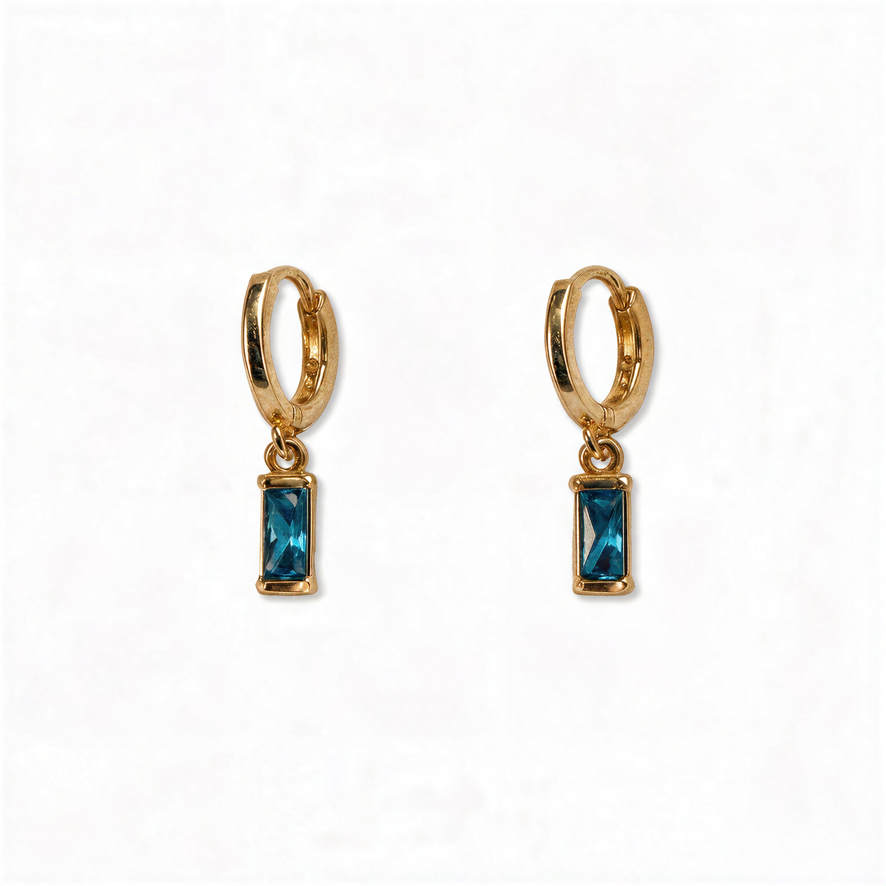 A pair of December Birthstone Aqua Cubic Zirconia Baguette Earrings by Skōsh Jewelry, featuring small gold huggie hoops, each with a rectangular blue baguette stone dangling below, displayed against a white background.