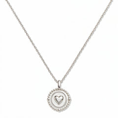 A Pass Love Along Heart Disc Pendant Necklace by Skōsh Jewelry, featuring a round medallion with a raised heart at its center framed by a beaded border on a fine sterling silver chain, displayed against a white background.