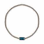 A December Birthstone Aqua Cubic Zirconia Baguette Bracelet by Skōsh Jewelry, featuring a strand of polished sterling silver beads on a stretch cord with a rectangular blue baguette stone at the center, displayed against a white background.