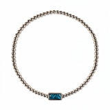 A December Birthstone Aqua Cubic Zirconia Baguette Bracelet by Skōsh Jewelry, featuring a strand of polished sterling silver beads on a stretch cord with a rectangular blue baguette stone at the center, displayed against a white background.