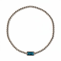 A December Birthstone Aqua Cubic Zirconia Baguette Bracelet by Skōsh Jewelry, featuring a strand of polished sterling silver beads on a stretch cord with a rectangular blue baguette stone at the center, displayed against a white background.