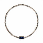 A September Birthstone Sapphire Cubic Zirconia Baguette Bracelet by Skōsh Jewelry, featuring a strand of polished sterling silver beads on a stretch cord with a rectangular deep blue baguette stone at the center, displayed against a white background.