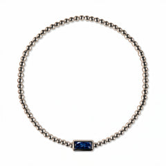 A September Birthstone Sapphire Cubic Zirconia Baguette Bracelet by Skōsh Jewelry, featuring a strand of polished sterling silver beads on a stretch cord with a rectangular deep blue baguette stone at the center, displayed against a white background.