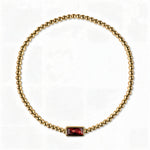 A January Birthstone Garnet Cubic Zirconia Baguette Bracelet by Skōsh Jewelry, featuring a strand of polished gold beads on a stretch cord with a rectangular deep red baguette stone at the center, displayed against a white background.
