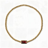 A January Birthstone Garnet Cubic Zirconia Baguette Bracelet by Skōsh Jewelry, featuring a strand of polished gold beads on a stretch cord with a rectangular deep red baguette stone at the center, displayed against a white background.