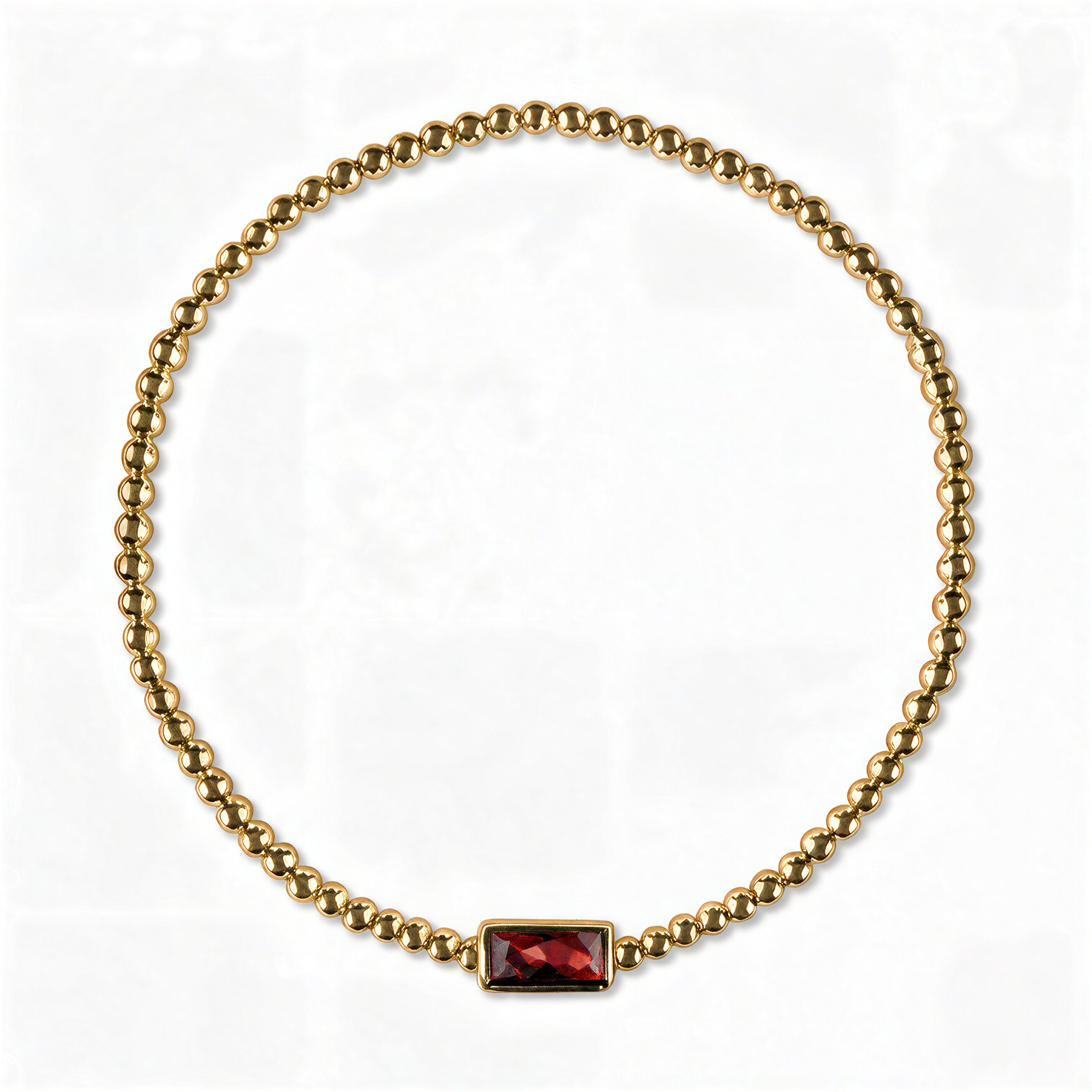 A January Birthstone Garnet Cubic Zirconia Baguette Bracelet by Skōsh Jewelry, featuring a strand of polished gold beads on a stretch cord with a rectangular deep red baguette stone at the center, displayed against a white background.