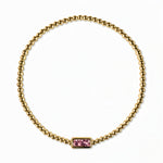 An October Birthstone Pink Cubic Zirconia Baguette Bracelet by Skōsh Jewelry, featuring a strand of polished gold beads on a stretch cord with a rectangular pink baguette stone at the center, displayed against a white background.