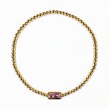 An October Birthstone Pink Cubic Zirconia Baguette Bracelet by Skōsh Jewelry, featuring a strand of polished gold beads on a stretch cord with a rectangular pink baguette stone at the center, displayed against a white background.